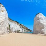 Spiaggia segreta in Puglia con sabbia dorata e acque cristalline, circondata da scogliere e vegetazione.